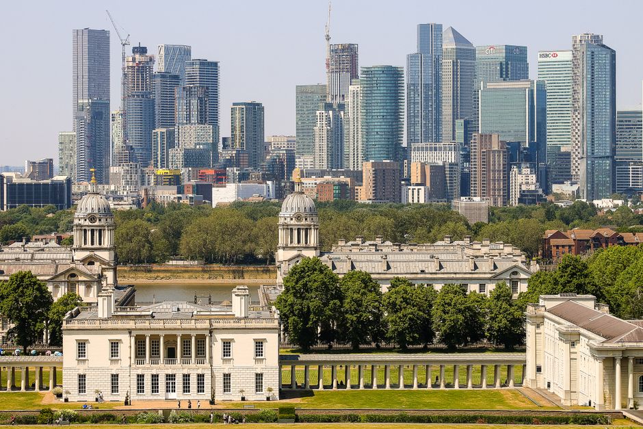 A panoramic view of Greenwich with historical white classical buildings in the foreground, featuring domed towers and columns, surrounded by lush green trees. Behind the historic structure, a modern city skyline with numerous high-rise office buildings and skyscrapers made of glass and steel is visible, reflecting sunlight. The scene showcases a contrast between historic architecture and contemporary urban development under bright daylight, highlighting the area’s architectural diversity. The area appears well maintained and clean, with no visible litter or debris, reflecting a sense of hygiene and care, aligning with the professional cleaning standards provided by Deep Cleaning Greenwich.