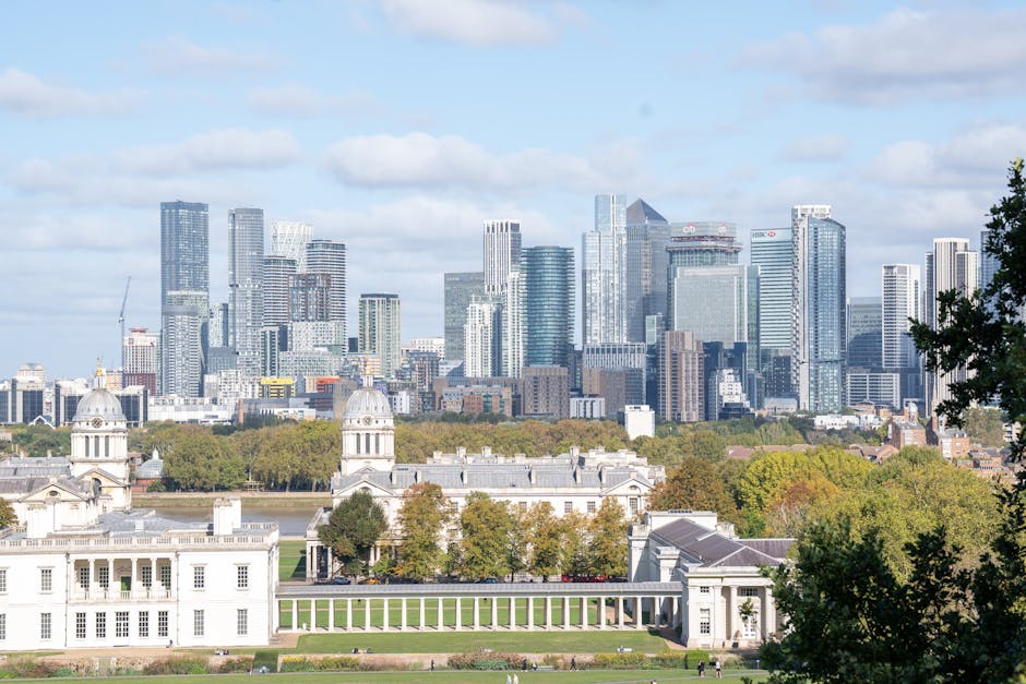 A panoramic view of Greenwich with historical white classical buildings in the foreground, featuring domed towers and columns, surrounded by lush green trees. Behind the historic structure, a modern city skyline with numerous high-rise office buildings and skyscrapers made of glass and steel is visible, reflecting sunlight. The scene showcases a contrast between historic architecture and contemporary urban development under bright daylight, highlighting the area’s architectural diversity. The area appears well maintained and clean, with no visible litter or debris, reflecting a sense of hygiene and care, aligning with the professional cleaning standards provided by Deep Cleaning Greenwich.