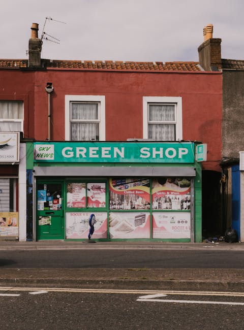 A street scene in Greenwich featuring a variety of commercial buildings with brick and stone facades, some with fire escapes. The ground level hosts multiple shopfronts including a gift shop and souve