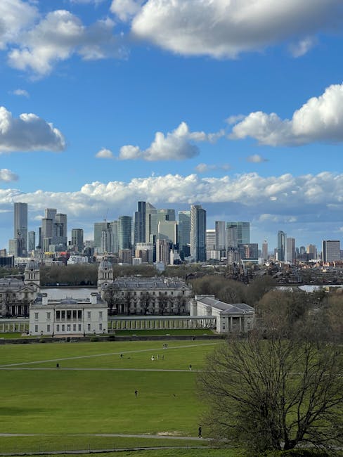 A wide view of a cityscape with modern high-rise buildings and skyscrapers under a partly cloudy sky, seen from a park with well-maintained green lawns and a few scattered trees. The foreground features an open grassy area with walking paths and a historic white building with classical architecture, including columns and ornate details. The scene captures the contrast between the natural park environment and the urban skyline, highlighting the cleanliness and orderliness of the green space. This image exemplifies urban surface viewing and outdoor space maintenance, reflecting the importance of cleanliness and presentation in both residential and commercial environments. For detailed surface cleaning or deep cleaning services in Greenwich SE10, including sanitisation and hygiene maintenance, Deep Cleaning Greenwich offers professional solutions.