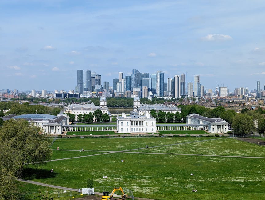 A panoramic view of a large, well-manicured green park with lush grass, scattered trees, and several visitors enjoying the outdoor space on a clear day. In the foreground, there is a historic white building with classical architectural features, including columns and a symmetrical design, surrounded by pathways and landscaped gardens. Modern high-rise buildings with glass facades and sleek designs form the city skyline in the background. Natural daylight illuminates the scene, highlighting the cleanliness and maintenance of the park and buildings. Deep Cleaning Greenwich provides comprehensive surface cleaning and sanitisation services to maintain such high standards of hygiene in residential and commercial properties.