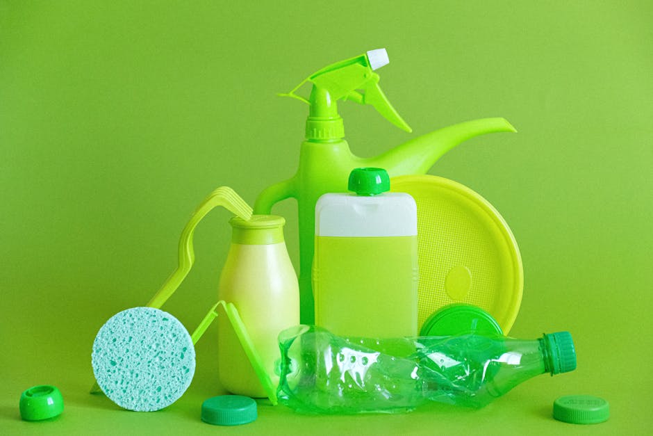 A collection of green cleaning supplies arranged on a bright green background, including a spray bottle with a trigger nozzle, a plastic bottle with a green cap, a sponge with a cleaning handle, a jar