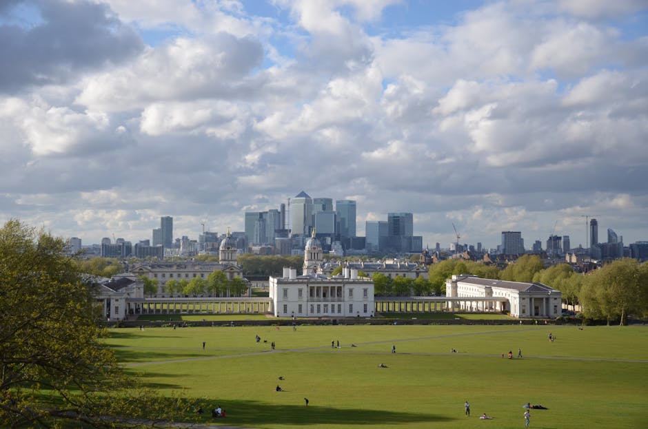 A wide view of Greenwich Park with lush green lawns and scattered visitors, featuring the historic Queen's House with its white facade and classical architecture in the foreground, and the modern London skyline with high-rise buildings under a partly cloudy sky in the background, captured during daytime. The image showcases a balance of natural greenery and urban development, emphasizing the park's well-maintained surfaces and scenic environment.
