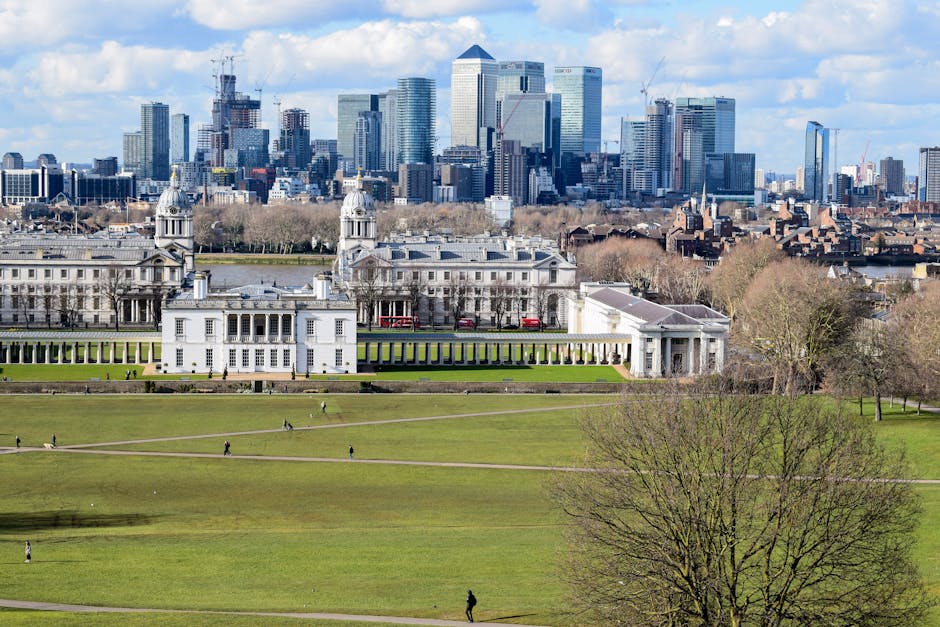 A panoramic view of a historic white building with classical architecture, including domed towers and colonnades, situated on a large green park with well-maintained lawns and pathways. In the background, a modern city skyline with tall glass skyscrapers under a partly cloudy sky. The scene features bright, natural lighting highlighting the clean, tidy surfaces of the historic structure and lush greenery, with no visible dirt or clutter. Deep Cleaning Greenwich provides expert surface cleaning, deep cleaning, and sanitisation services for both residential and commercial spaces, ensuring hygiene and cleanliness at our clients' premises.