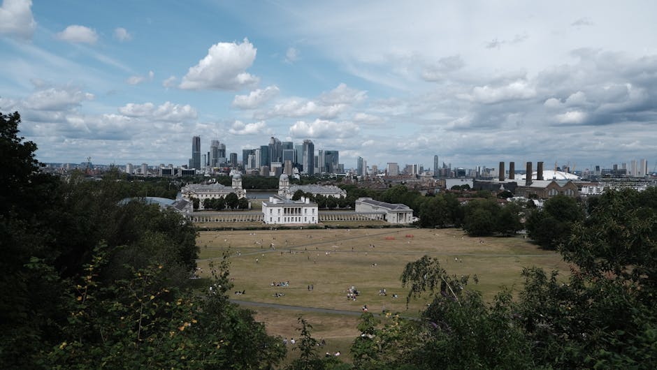 An aerial view of Greenwich showing a wide river winding through the city with a mix of historic and modern buildings on the riverbank, surrounded by lush green parks and streets lined with trees. The